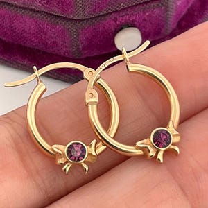 May include: A pair of gold hoop earrings, each featuring a small, round, purple gemstone set within a decorative bow. The earrings are displayed against a blurred background.