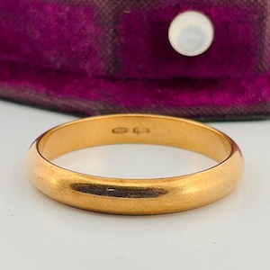 May include: A polished gold band ring with a smooth, rounded surface. The interior of the band features small, engraved hallmarks indicating its metal purity. The ring is a simple, solid gold circle with a reflective, metallic finish throughout.