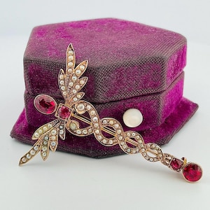 May include: A gold brooch with a swirling design, decorated with small pearls and red gemstones. The brooch features a stylized leaf and a large red gemstone. It rests on a purple velvet box.