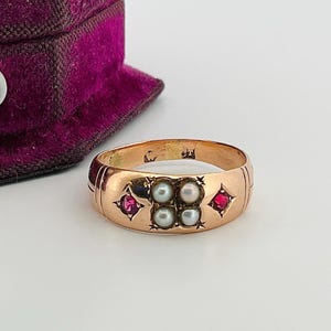 May include: A gold ring featuring four small pearls at the centre, with two red gemstones on either side. The ring has an etched band design. A purple velvet jewellery box is partially visible in the background.