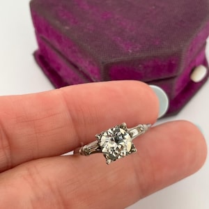 Peut inclure: Une bague argentée dotée d'une pierre ronde transparente proéminente montée sur quatre griffes, flanquée de deux petites pierres taille baguette de chaque côté de l'anneau, présentée avec une boîte à bijoux hexagonale en velours violet.