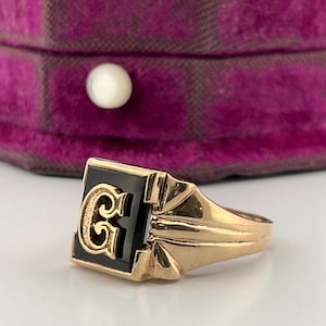 Puede incluir: Un anillo dorado con un cuadrado de ónix negro con una letra "G" dorada. El anillo se encuentra sobre un fondo borroso de una caja de terciopelo púrpura con un botón blanco perlado.