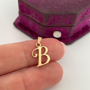 Puede incluir: Un colgante dorado con la forma de la letra "B" con un adorno decorativo. El colgante se muestra delante de un joyero de terciopelo morado oscuro.