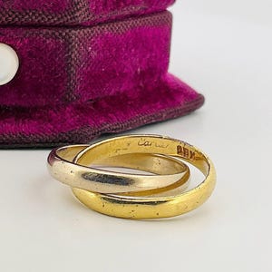 May include: Three-band gold ring with the word "Cartier" and "ABM" engraved on the surface. The ring is set against a purple velvet jewelry box.