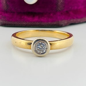 May include: A gold ring featuring a single round brilliant-cut diamond set in a smooth, circular white metal bezel. The band is crafted from polished yellow gold, creating a two-tone appearance with the central stone setting.