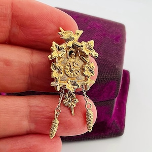 以下が含まれることがあります： 金色のカッコウ時計ペンダント。花と鳥の複雑なディテールが特徴です。時計は丸い文字盤、上部の鳥、ぶら下がっている松ぼっくりのチャームが特徴です。ペンダントは紫色のベルベットの背景に配置されています。