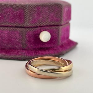 May include: A tri-color rolling ring featuring three interlocking bands in yellow, rose, and white gold. The smooth, polished metal bands are intertwined to form a single circular piece of jewelry, showcasing a classic design with metallic tones.