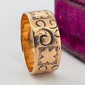May include: A gold band ring featuring an engraved pattern of alternating four-leaf clover shapes set within square borders and swirling scroll motifs. The metal surface has a polished finish with recessed areas that highlight the intricate design.