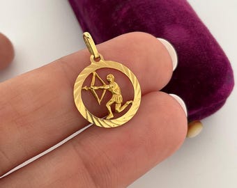 May include: A gold-colored pendant featuring the Sagittarius zodiac symbol. The pendant depicts an archer with a bow and arrow within a circular frame. The pendant is suspended from a small loop, ready to be worn. The background includes a purple velvet surface.