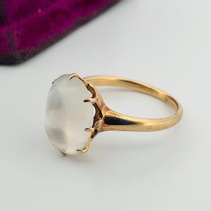 Pu&ograve; includere: Un anello d'oro con una grande pietra ovale bianco latte. La fascia e l'incastonatura dell'anello sono dorate e la pietra &egrave; tenuta da delicate griffe. L'anello &egrave; posto su uno sfondo bianco.