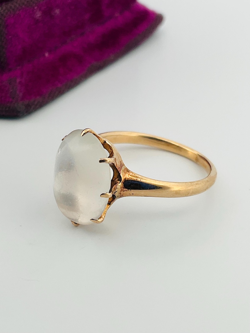 Pu&ograve; includere: Un anello d'oro con una grande pietra ovale bianco latte. La fascia e l'incastonatura dell'anello sono dorate e la pietra &egrave; tenuta da delicate griffe. L'anello &egrave; posto su uno sfondo bianco.