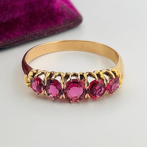 Peut inclure: Une bague en or ornée de cinq pierres précieuses roses vives. Les pierres sont disposées en rang, créant une bande de couleur continue. La bague a un design classique avec une bande en or poli.