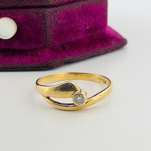 Op de afbeelding: Een gouden ring met een kleine, heldere edelsteen in een gebogen ontwerp. De ring ligt op een wit oppervlak, met een paarse fluwelen sieradendoos op de achtergrond.