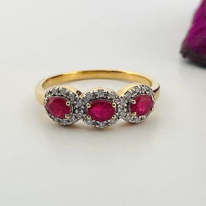 May include: A gold ring featuring three oval-cut, vibrant red gemstones, each surrounded by a halo of smaller, clear stones. The ring's band is a polished gold color, creating a classic and elegant design.