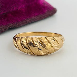 May include: A gold band ring featuring a domed profile with a textured surface. The ring displays alternating diagonal patterns of polished, reflective gold and matte, brushed gold finishes across the entire outer circumference of the band.