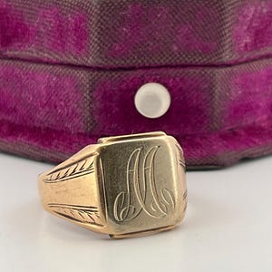 May include: A gold signet ring featuring a rectangular face with an engraved script letter M. The band displays decorative etched leaf patterns on both sides, showcasing a polished metallic finish and a classic, structured design.