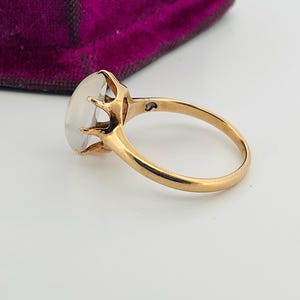 Pu&ograve; includere: Un anello d'oro con una grande gemma ovale e traslucida. La fascia dell'anello &egrave; in oro lucido e la gemma &egrave; tenuta in posizione da delicate griffe. L'anello &egrave; posto su uno sfondo bianco, con uno scorcio di un cofanetto di velluto viola.