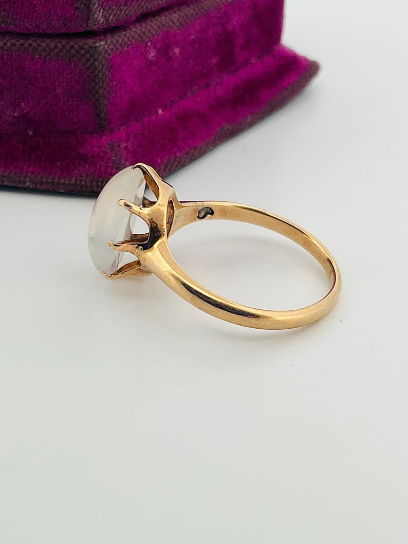 Pu&ograve; includere: Un anello d'oro con una grande gemma ovale e traslucida. La fascia dell'anello &egrave; in oro lucido e la gemma &egrave; tenuta in posizione da delicate griffe. L'anello &egrave; posto su uno sfondo bianco, con uno scorcio di un cofanetto di velluto viola.