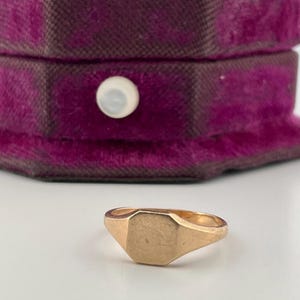 Puede incluir: Un anillo de sello de oro con una cara octogonal lisa y plana, colocado frente a un joyero de terciopelo morado oscuro que presenta un pequeño botón de cierre redondo y blanco en su parte frontal.
