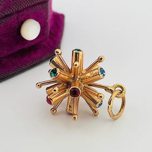 May include: A gold-toned charm with a starburst design. The charm features a central red gemstone surrounded by radiating gold rods, each tipped with a small, colorful gem. The gems are red, green, and blue. A small gold ring is attached.