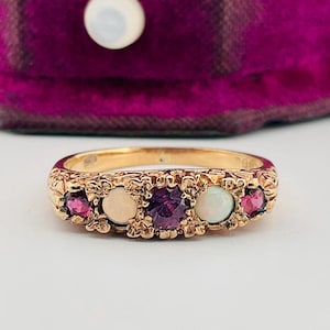 May include: A gold ring featuring a central round purple gemstone flanked by two round white opals and two smaller round dark pink garnets. The band has a textured, ornate metal finish with five distinct stone settings arranged horizontally.