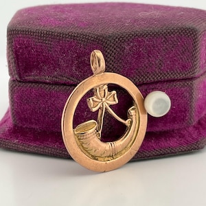 May include: A gold-coloured pendant with a hunting horn and a cross design within a circular frame. The pendant is set against a dark purple velvet background. A small, round, white pearl is visible.