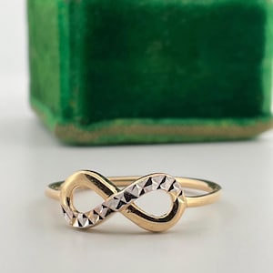 May include: A gold-coloured ring with an infinity symbol design. The infinity symbol has a textured, diamond-cut pattern in a contrasting silver colour. The ring is set against a blurred green background.