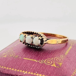 May include: A gold ring with three opal gemstones. The opals are set in a decorative band. The ring is displayed on a purple surface with gold accents. The ring's design is vintage.