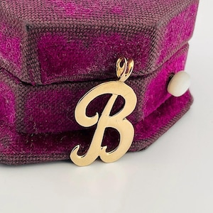 Puede incluir: Un colgante dorado con forma de la letra "B" en fuente cursiva. El colgante se muestra sobre una caja de joyería morada y marrón con un cierre de botón blanco.