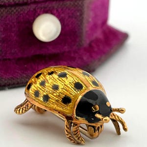 May include: A gold-tone beetle brooch featuring a textured yellow-gold body with black enamel spots and a smooth black enamel head. The insect-shaped pin has detailed legs and antennae, showcasing a metallic finish throughout the design.