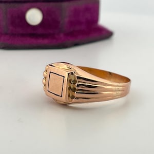 May include: A gold signet ring with a square face and a ridged band. The ring's face has a rectangular design. The ring is set against a white surface with a purple velvet box in the background.