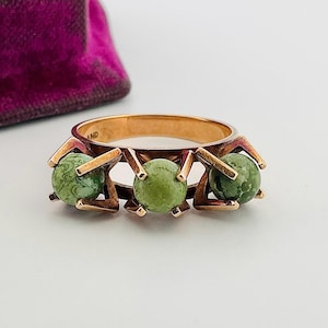 May include: A gold-toned ring featuring three round, green gemstones. The gemstones are set within a geometric design. The ring is displayed against a white background, with a glimpse of a purple velvet box.
