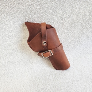 May include: A brown leather holster with a belt loop and buckle. The holster is designed to hold a handgun and is made of a sturdy, durable material. The holster is a classic design, perfect for Western-themed costumes or historical reenactments.