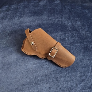 May include: A tan leather holster featuring a curved edge design, a single strap with a silver-toned metal snap button closure, and an adjustable strap with a silver-toned metal buckle for securing the contents within the main compartment.