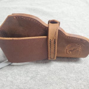 May include: A brown leather holster with a stitched design and a metal buckle. The holster has a logo that says "Horse Leather" with a star and a horse silhouette.