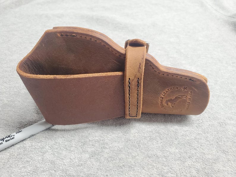 May include: A brown leather holster with a stitched design and a metal buckle. The holster has a logo that says "Horse Leather" with a star and a horse silhouette.