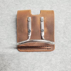 May include: A brown leather hammer holder with two metal loops and a metal ring. The holder has a stitched edge.
