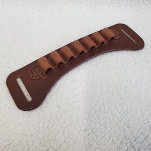 May include: A brown leather cartridge holder featuring a curved, elongated base with six stitched loops for holding individual rounds. The piece includes a rectangular slot at each end for attachment, showcasing visible stitching and a smooth finish.