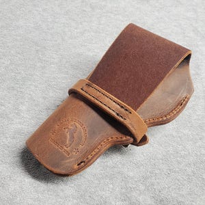 May include: A brown leather holster with a stitched flap and a logo that reads "The Old West Holsters Made in USA".