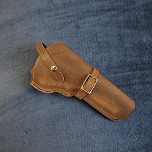 May include: A brown leather holster featuring a curved top edge, a single strap secured with a metallic snap button, and a secondary adjustable strap across the body fastened with a rectangular silver-toned metal buckle for secure closure.
