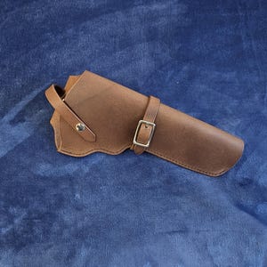 May include: A brown leather holster featuring a smooth finish, a curved edge design, a single silver-toned metal snap button closure on the side strap, and a thin adjustable strap secured with a rectangular silver-toned metal buckle.