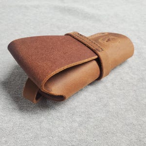 May include: A brown leather case with a flap closure and a stitched strap. The case is folded closed and lying on a light grey surface.
