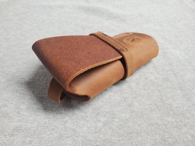 May include: A brown leather case with a flap closure and a stitched strap. The case is folded closed and lying on a light grey surface.