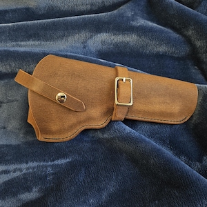 May include: A tan leather holster featuring a single strap with a metallic snap button closure and a secondary adjustable strap with a rectangular metal buckle. The holster is constructed with visible dark stitching along the edges.