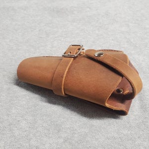 May include: A brown leather holster with a silver buckle and a strap. The holster is designed to hold a firearm.