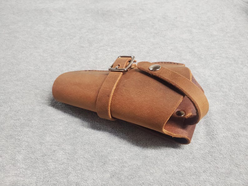 May include: A brown leather holster with a silver buckle and a strap. The holster is designed to hold a firearm.