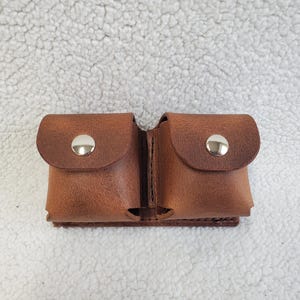 May include: A brown leather pouch with two compartments. Each compartment has a rounded flap secured by a silver snap. The pouch is designed to hold small items and is made of textured leather.