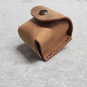 May include: A small, brown leather box with a flap closure and a black button. The box is stitched around the edges and has a worn, rustic look.