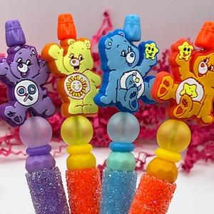 Op de afbeelding: Vier pennen met Care Bear-thema, elk met een ander personage in paars, geel, blauw en oranje. De pennen hebben glinsterende, getextureerde barrels en zijn bekroond met een kleine, gekleurde dop.