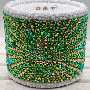 May include: A cylindrical container covered in small, reflective green and gold rhinestones arranged in a radiating pattern. The top and bottom edges are bordered by a thick band of small, matte white spherical beads, creating a textured finish.